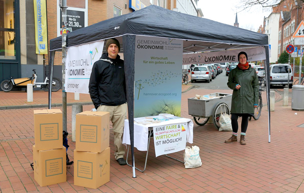 Zu sehen ist der Info-Stand der Gemeinwohl-Ökonomie Hannover auf dem Lindener Marktplatz. Es ist ein Pavillon aufgebaut, der von mehreren Seiten bedruckt ist. Auf einem Tisch liegt allerlei Info-Material, und zwei fröhlich blickende Mitglieder unserer Regionalgruppe betreuen den Stand.