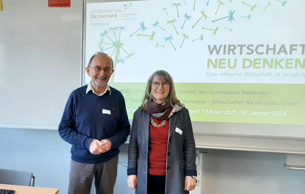 Fachtag Gymnasium Mellendorf: Die beiden Referent:innen der Regionalgruppe Hannover stehen vor einer Projektion des GWÖ-Logos, der Beschriftung 'Wirtschaft neu denken', und lächen offen in die Kamera.